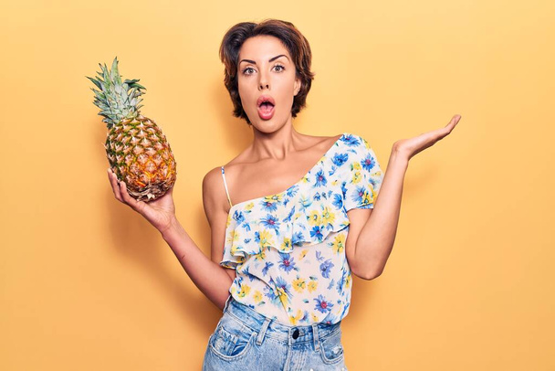 Young beautiful woman holding pineapple scared and amazed with open mouth for surprise, disbelief face  - Фото, изображение