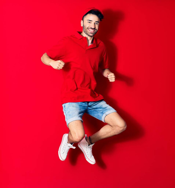 Young handsome deliveryman wearing cap smiling happy. Jumping with smile on face over isolated red background - Foto, afbeelding