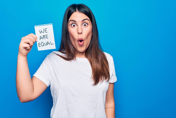 Young beautiful brunette woman asking for equality holding paper with we are equal message scared and amazed with open mouth for surprise, disbelief face - Photo, Image