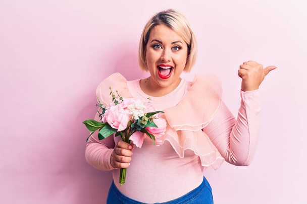Schöne blonde Plus-Size-Frau mit einem Strauß rosa Blumen über isoliertem Hintergrund, der den Daumen zur Seite zeigt und glücklich mit offenem Mund lächelt - Foto, Bild