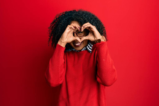 Mooie Afrikaanse amerikaanse vrouw met afro haar dragen trui en bril doen hart vorm met de hand en vingers glimlachend kijken door middel van teken  - Foto, afbeelding