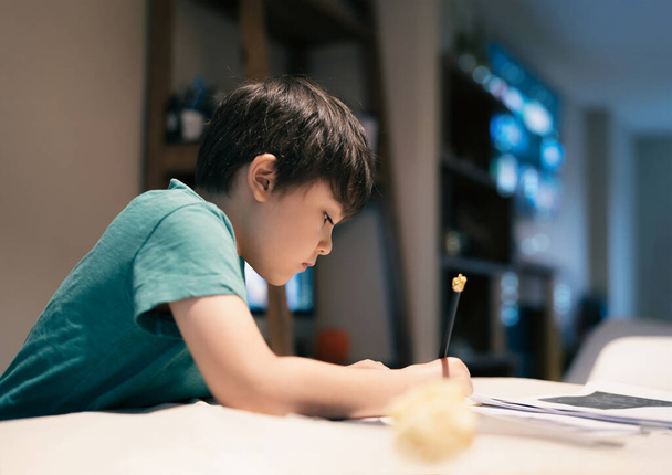 Portret chłopca ze szkoły siedzącego na stole odrabiającego lekcje, dziecko trzymające ołówek do pisania, chłopiec rysujący na białym papierze na stole, szkoła podstawowa i koncepcja wychowania domowego - Zdjęcie, obraz