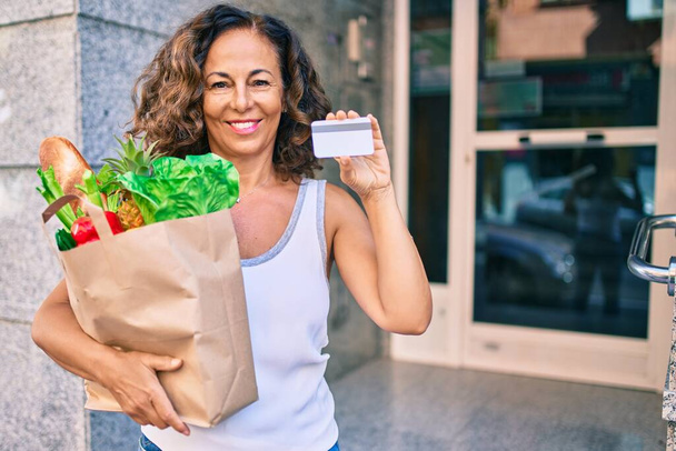 Orta yaşlı İspanyol kadın, elinde alışveriş poşeti ve kredi kartıyla şehirde mutlu bir şekilde gülümsüyor.. - Fotoğraf, Görsel