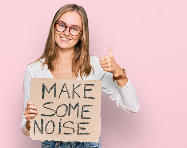 Beautiful blonde woman holding make some noise banner smiling happy and positive, thumb up doing excellent and approval sign  - Photo, Image
