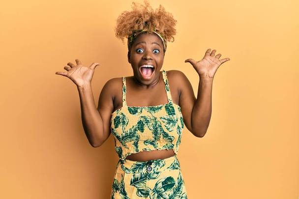 Mujer africana joven con pelo afro vistiendo vestido de verano celebrando loco y sorprendido por el éxito con los brazos levantados y los ojos abiertos gritando emocionado. concepto ganador  - Foto, imagen