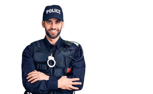 Młody przystojniak w brodzie w mundurze policyjnym uśmiechnięty ze skrzyżowanymi rękami patrzący w kamerę. osoba pozytywna.  - Zdjęcie, obraz
