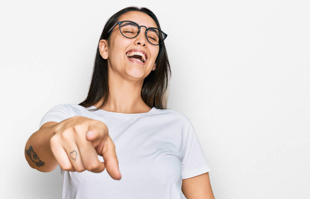 Mujer hispana joven vistiendo camiseta blanca casual riéndose de ti, señalando con el dedo a la cámara con la mano sobre el cuerpo, expresión de vergüenza  - Foto, imagen