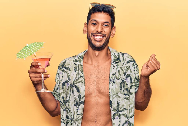 Joven hombre latino con camisa de verano sosteniendo cóctel gritando orgulloso, celebrando la victoria y el éxito muy emocionado con el brazo levantado  - Foto, Imagen