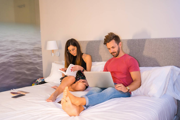 A Caucasian couple lounging on the hotel bed and looking at a computer and a book. Summer lifestyle - Fotoğraf, Görsel