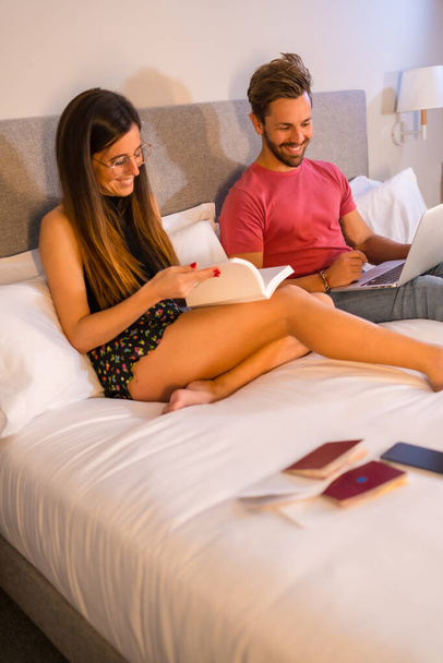A handsome Caucasian couple lounging on the hotel bed and looking at a computer and a book. Summer lifestyle - Foto, Imagem