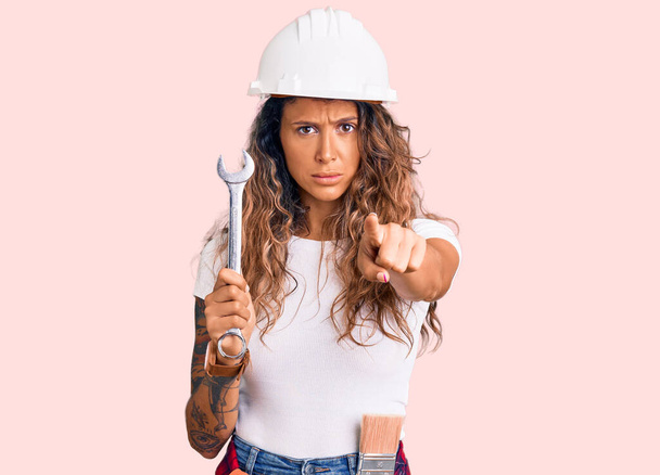 Young hispanic woman with tattoo wearing hardhat and builder clothes holding wrench pointing with finger to the camera and to you, confident gesture looking serious  - Photo, Image