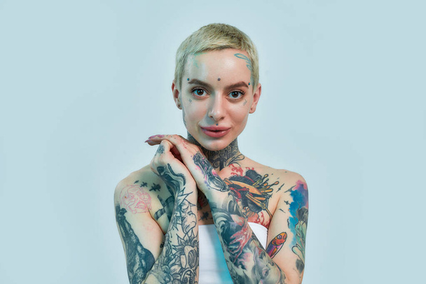 Tattoo and piercing. A tattooed and pierced white girl wearing denim overall standing and looking into a camera holding her hands together near her face and smiling - Photo, image