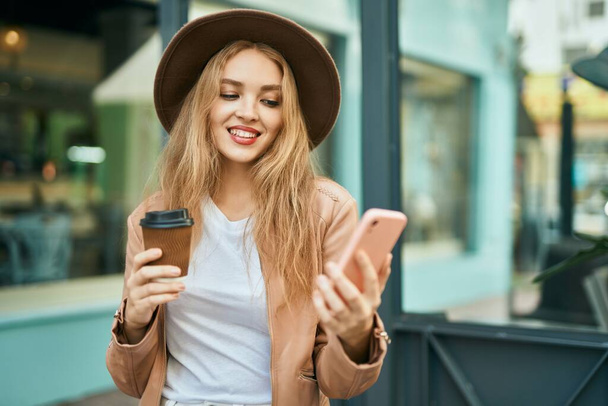 Jovencita rubia usando smartphone y tomando café en la ciudad. - Foto, Imagen