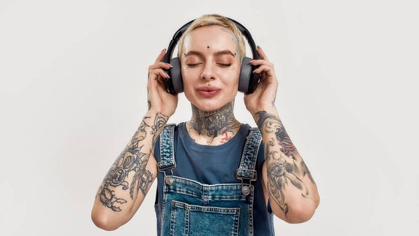 Listening to music. A young white pierced and tattoed woman wearing a denim overall listening to music with her big earphones with her eyes closed - Valokuva, kuva