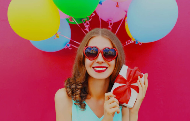 Portret uśmiechniętej młodej kobiety z pudełkiem prezentów i kolorowymi balonami na różowym tle - Zdjęcie, obraz