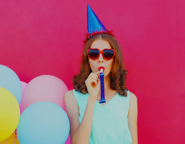 Portrait of cheerful young woman in birthday cap with colorful balloons on a pink background - Photo, image