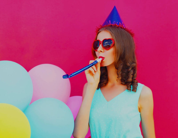 Portrait of cheerful young woman in birthday cap with colorful balloons on a pink background - Fotó, kép