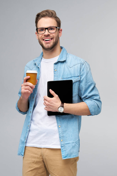 Happy young man wearing jeans shirt standing and using tablet pc pad and holding cup of coffee to go standing over studio grey background - Foto, afbeelding