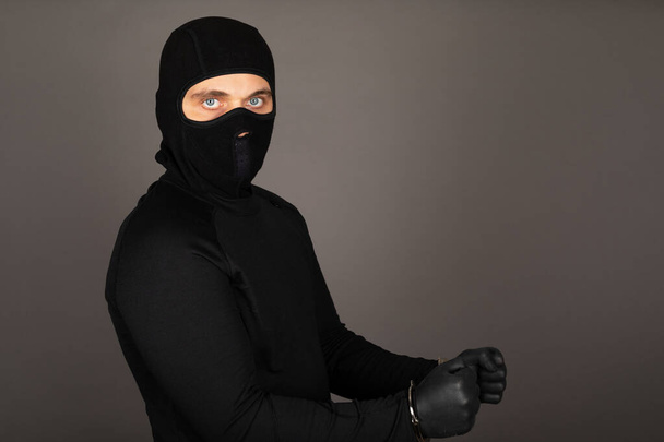 Picture of young man with black mask and outfit suspect of a robbery, wearing handcuffs in front of grey background - Photo, Image