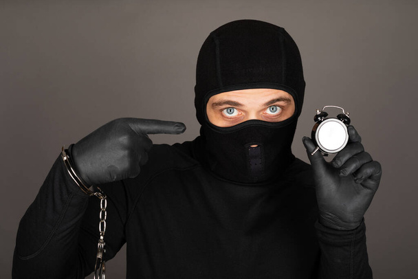 Picture of young man with black mask and outfit suspect of a robbery, wearing handcuffs in front of grey background - Foto, imagen