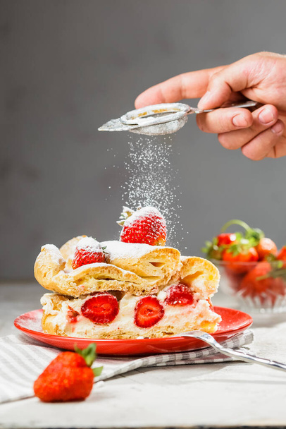 A piece of strawberry cake on a red plate. Man's hand with sieve icing sugar powder from above. Grey background, vertical composition - Photo, image