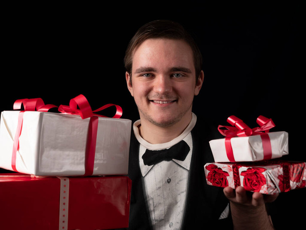 Ένας νεαρός με μαύρη στολή κρατά κόκκινα και λευκά δώρα στα χέρια του σε μαύρο φόντο. - Φωτογραφία, εικόνα