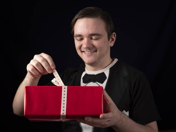 Junger Mann im schwarzen T-Shirt-Anzug öffnet ein rotes Geschenk auf schwarzem Hintergrund - Foto, Bild