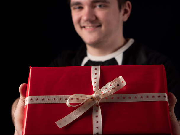 Ένας νεαρός με μαύρη στολή απλώνει το χέρι του με κόκκινο δώρο σε μαύρο φόντο. - Φωτογραφία, εικόνα