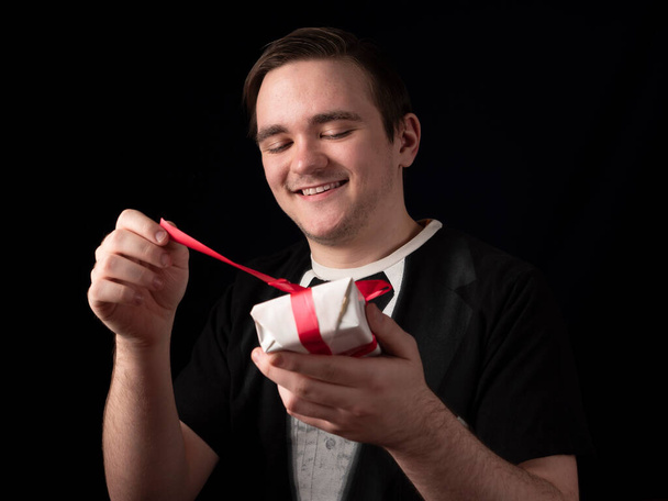young guy in a black t-shirt suit opens a white gift with a smile on a black background - Photo, Image