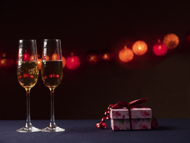 Два бокала шампанского и розовый подарок с красной лентой на черном фоне со светом - Фото, изображение