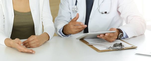 Doctor and patient discussing medical exam results while sitting at the desk in sunny clinic, close-up. Male physician using tablet computer for filling up medication history record. - Foto, Bild