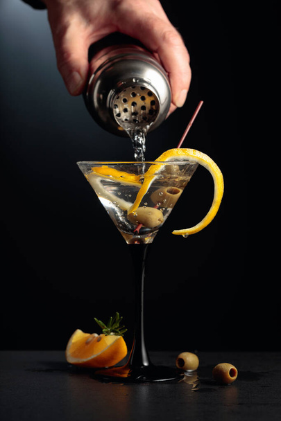 Barman giet cocktail uit shaker. Droge martini met citroenschil en groene olijven op een zwarte achtergrond. - Foto, afbeelding