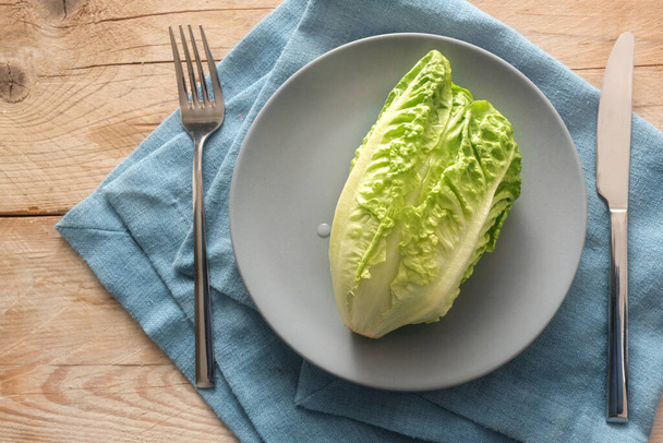 Fresh green lettuce or baby cos on a gray plate with cutlery on a blue napkin and rustic wooden planks, concept for a healthy vegetarian diet to lose weight, copy space, high angle view from above - Фото, изображение