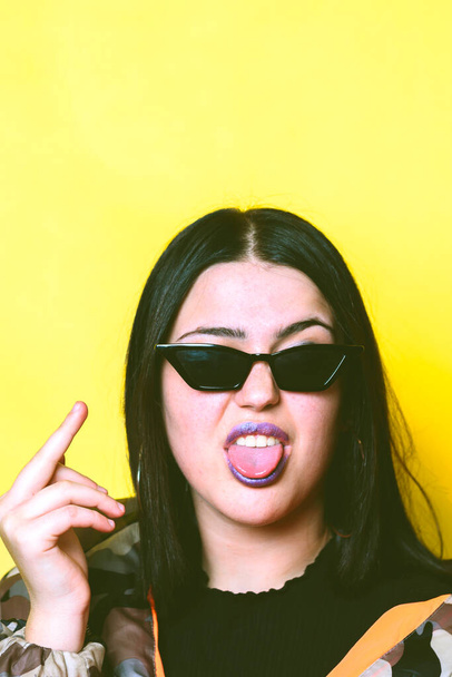Teenager with sunglasses and yellow background sticking out his tongue and reading the middle finger. - Foto, immagini
