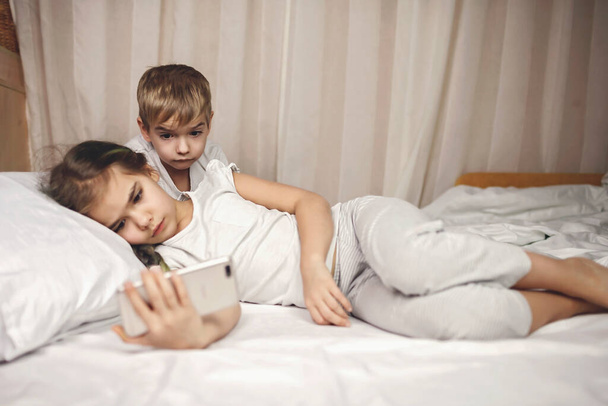 Deux frères et sœurs regardant la vidéo dans un appareil électronique mobile dans le lit, dépendance gadget, mode de vie - Photo, image
