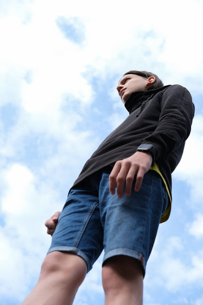 teenager boy wearing black hoodie and shorts. low angle view. boy standing over a sky background. unusual trendy shot. selective focus. vertical size. - Foto, Imagem