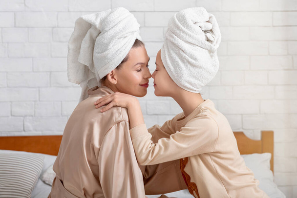 Zijaanzicht van jonge moeder en haar tienerdochter in badhanddoeken aanraken neuzen op bed thuis - Foto, afbeelding