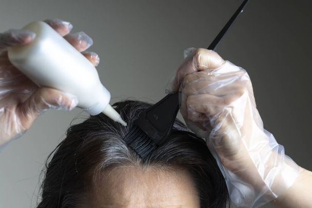 Closeup woman hands dyeing hair using black brush. Middle age woman colouring dark hair with gray roots at home - Photo, Image