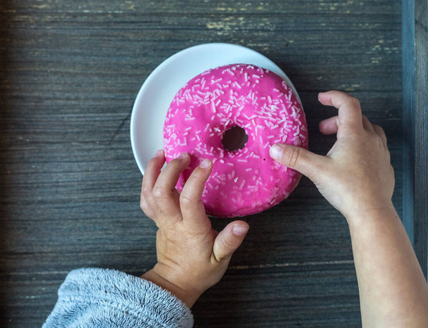 Baby takes bright pink donut from white plate. Childs hands. Grey wooden background. Sweets for kids breakfast. Traditional American meal. Top view to a table with round objects. Gluten products - Фото, изображение