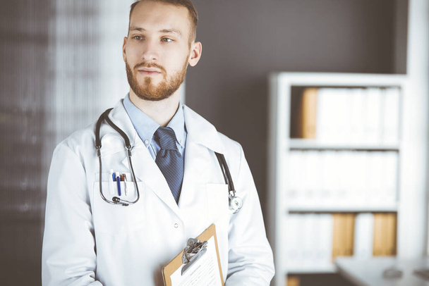 Red-bearded doctor standing straight in clinic near his working place. Portrait of physician. Medicine and healthcare concept - Photo, image