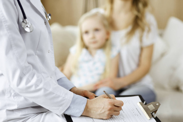 Doctor and patient. Pediatrician using clipboard while examining little girl with her mother at home. Happy cute caucasian child at medical exam. Medicine concept - Foto, imagen