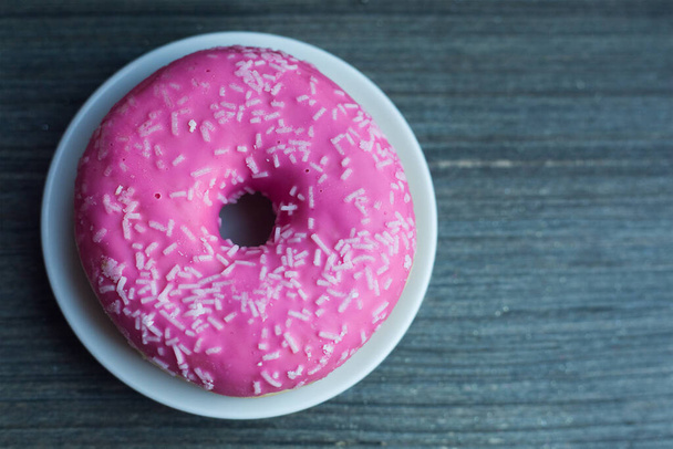 Top wetteifern zu leuchtend rosa Donut auf weißem runden Teller und gealtertem hölzernem grauen Hintergrund. Isolierte Objekte aus nächster Nähe. Traditionelle amerikanische Süßigkeiten zum Frühstück. Kopierraum. Weiße Zuckerdekoration obendrauf.  - Foto, Bild
