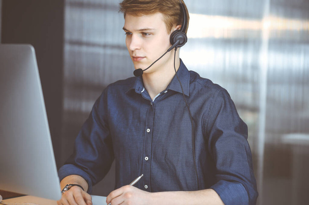 Young blond businessman using headset and computer at work. Startup business means working hard and out of time for success achievement - 写真・画像