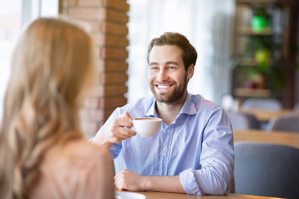 Улыбающийся парень тысячелетия пьет кофе со своей девушкой в кафе, весело разговаривает за завтраком - Фото, изображение