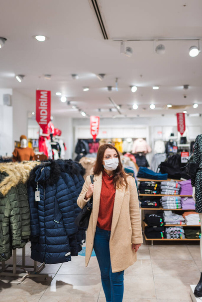 Beautiful girl wearing protective medical mask and fashionable clothes looks at clothes at store. New normal lifestyle concept. - Photo, Image