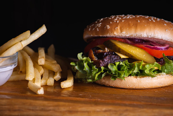 hamburger con fette al forno di carne fritta, pomodori, cipolle, formaggio, lattuga e patatine fritte su una superficie di legno isolata su fondo scuro. Il concetto di cibo delizioso - Foto, immagini