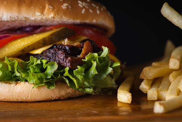 hamburger con fette al forno di carne fritta, pomodori, cipolle, formaggio, lattuga e patatine fritte su una superficie di legno isolata su fondo scuro. Il concetto di cibo delizioso - Foto, immagini