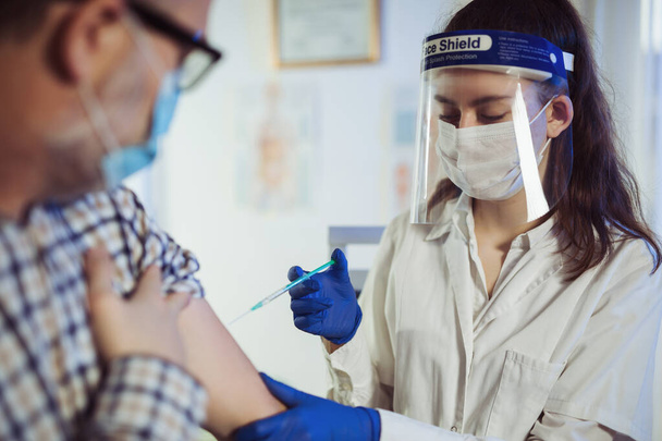 Cerrahi maskeli ve yüzlü genç bir hemşire yaşlı adama iğne yapıyor. Covid-19 veya koronavirüs aşısı - Fotoğraf, Görsel