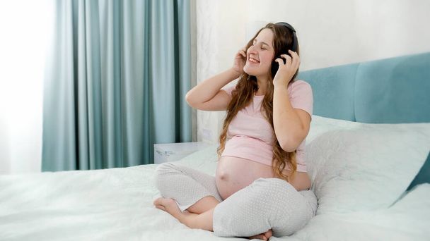 Feliz mujer embarazada sonriente en pijama escuchando música con auriculares en la cama por la mañana - Foto, Imagen