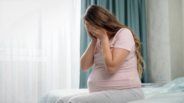 Droevig huilende zwangere vrouw die lijdt aan depressie zittend op bed en haar hoofd vasthoudend. Concept van maternale en zwangerschapsdepressie. - Foto, afbeelding